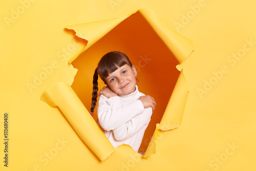 Indoor shot of egoistic little girl with braids wearing white turtleneck posing in torn hole of yellow paper wall, standing embracing her body, expressing love to herself.