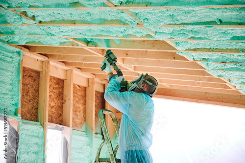 Male builder insulating wooden frame house. Man worker spraying polyurethane foam inside of future cottage, using plural component gun. Construction and insulation concept.