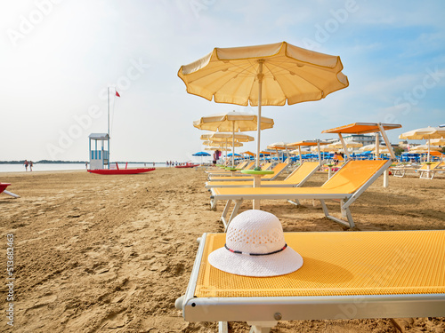 Fototapeta Naklejka Na Ścianę i Meble -  White straw hat on a beach bed with colorful umbrellas on a sunny summer day. In the background a lifeboat alongside a lifeguard tower on the shore of the Adriatic Sea, Italy.