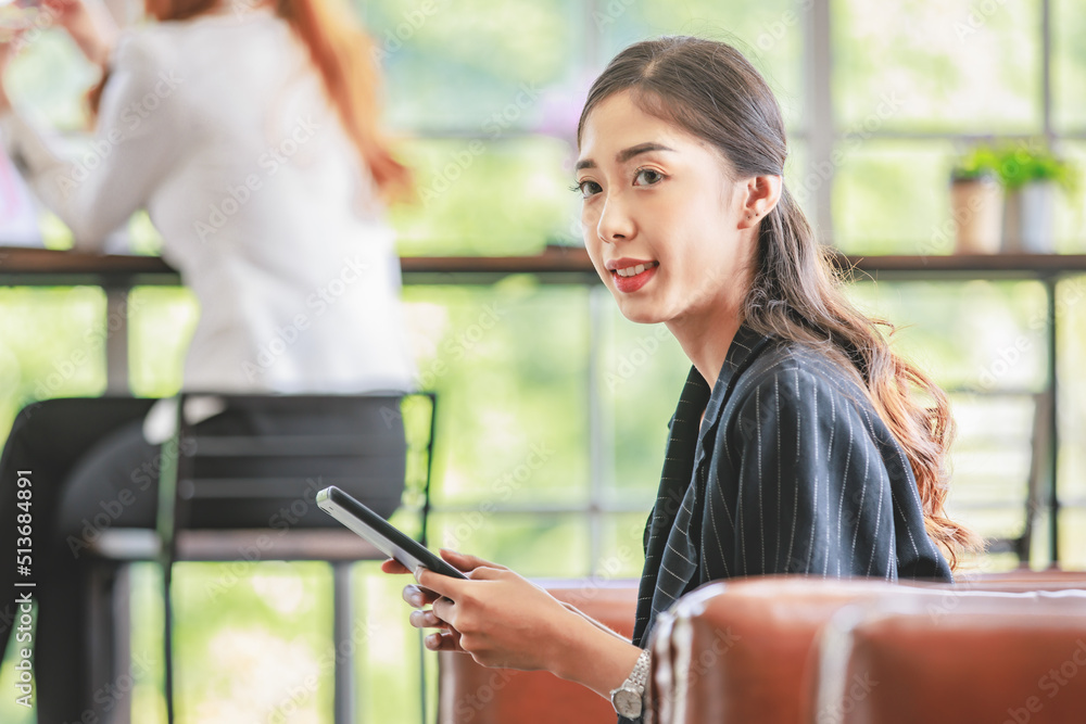 Asian young pretty professional successful female businesswoman sitting on sofa checking customer email via tablet when colleague using laptop computer working on counter table in company living room Asian young pretty professional successful female businesswoman sitting on sofa checking customer email via tablet when colleague using laptop computer working on counter table in company living room