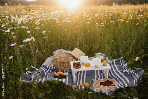 Beautiful summer picnic with fresh herbal chamomile tea, croissants and pie on flower meadow at sunset.