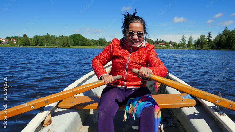 aktive asiatische Touristin rudert bei Wind über See in Bayern zu grünem Wald bei sonnigem blauen Himmel