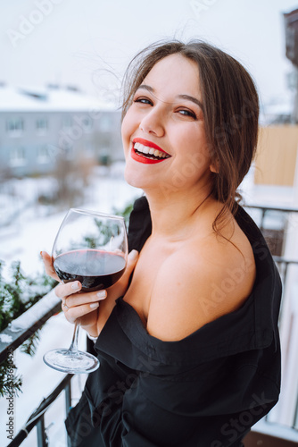 Young happy laughing woman wearing black shirt with bare shoulder, standing on balcony, holding glass with red wine.