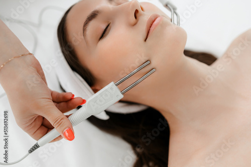 A young girl with a white bandage on her head lies in the cosmetologist's office, view from above. the hands of a female cosmetologist perform a microcurrent facial treatment with the machine. spa