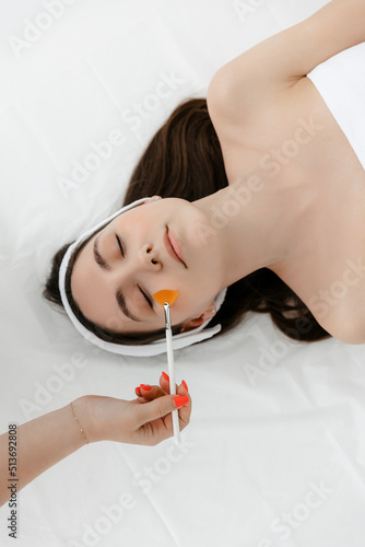 A beautiful young Caucasian girl with a white bandage on her head is lying in the cosmetologist's office, top view. The beautician applies a liquid brush to her face. chemical or acid peel, moisturize