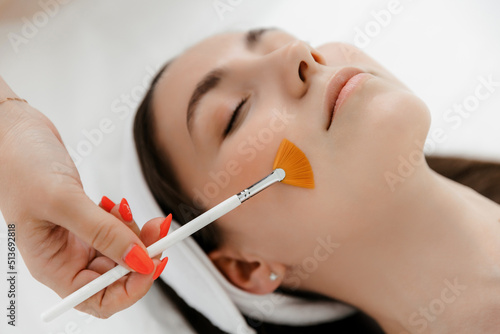 A beautiful young Caucasian girl with a white bandage on her head is lying in the cosmetologist's office, top view. The beautician applies a liquid brush to her face. chemical or acid peel, moisturize