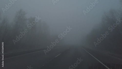 The car is driving along an asphalt road in the fog along the forest. Car are passing towards us. View through the windshield