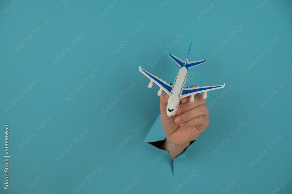 A female hand sticking out of a hole from a blue background holds a model of an airplane. A female hand sticking out of a hole from a blue background holds a model of an airplane.