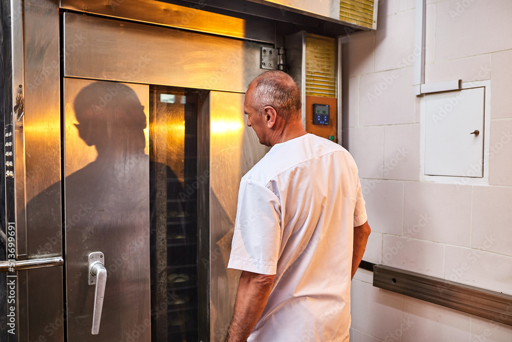 Professional baker in uniform stands near industrial oven in a bakery for making bread Professional baker in uniform stands near industrial oven in a bakery for making bread