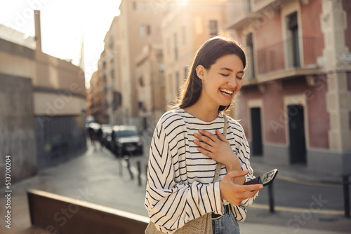 Positive young caucasian girl laughs from watching video on phone standing outdoors. Brunette with closed eyes wears white sweatshirt. Mood, lifestyle, concept.