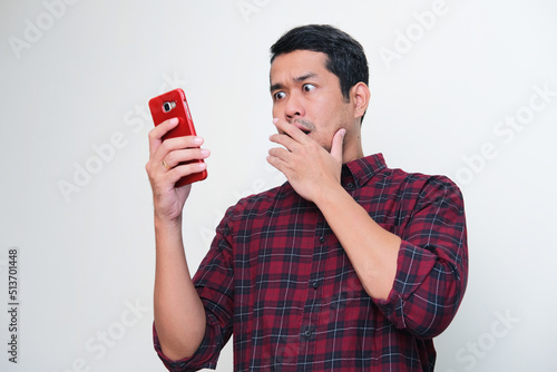 Adult Asian man showing shocked expression when looking to his mobile phone photo