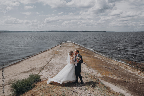 Groom in a black suit with a black bow tie with a beautiful bride in a white long dress embrace and kiss  holding hands near the river. Wedding portrait of newlyweds.