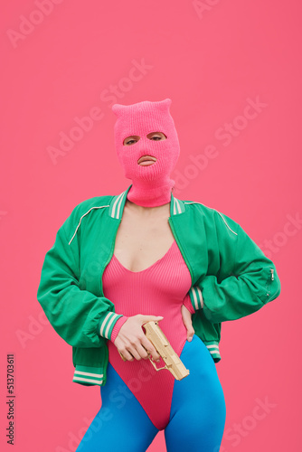 Portrait of fashion model in pink balaclava and body posing with gun against pink background expressing protest photo