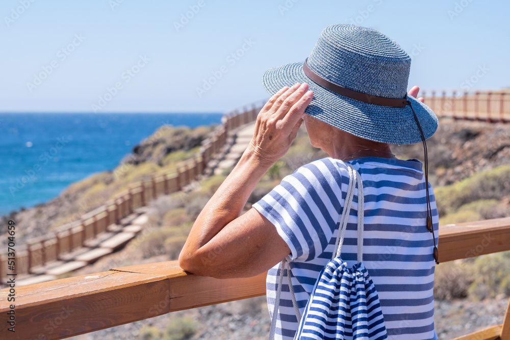 Rear view of senior caucasian woman holding her hat while looking at the horizon over water enjoying summer holidays