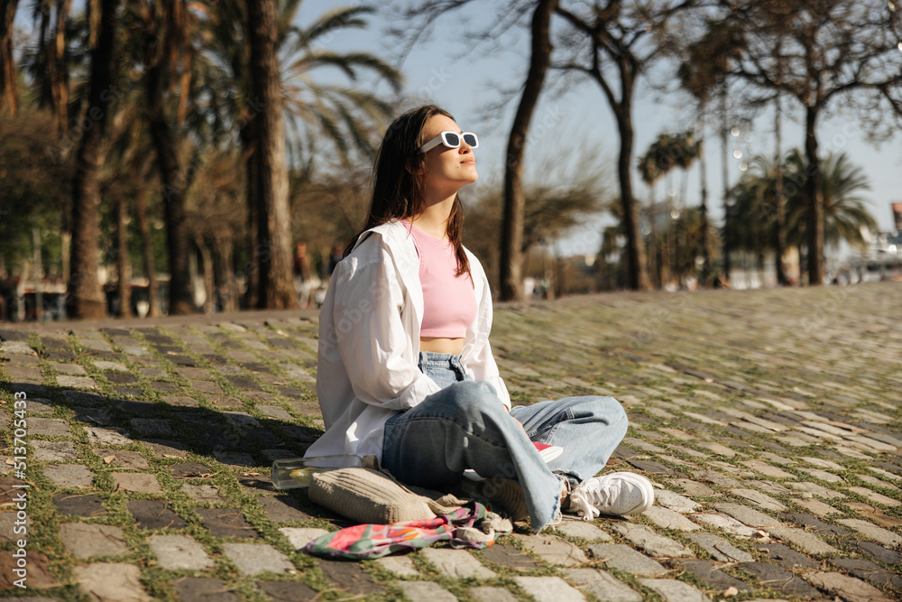 Pretty young caucasian girl is resting sitting on ground in sunny weather outdoors. Brunette wears sunglasses, casual clothes. Lifestyle concept Pretty young caucasian girl is resting sitting on ground in sunny weather outdoors. Brunette wears sunglasses, casual clothes. Lifestyle concept