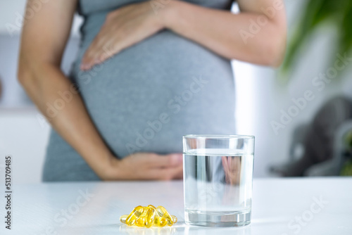Vitamin D capsules and glass of water and pregnant woman at home interior. Healthy fatty acids nutritional supplement for prenatal support. Omega, DHA, vitamin D, fish oil for healthy pregnancy.