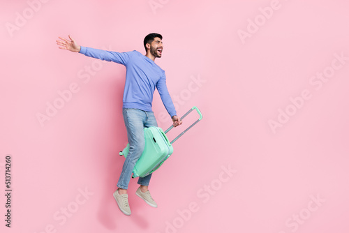 Full length photo of excited pretty arabian man wear long sleeve shirt riding suitcase jumping empty space isolated pink color background photo