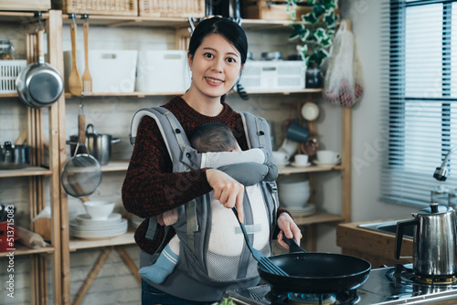 portrait cheerful asian housewife holding a spatula with infant child in the carrier is smiling at camera while preparing meal in the morning at home kitchen.