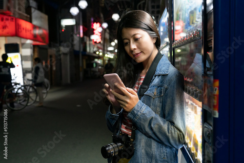 pretty asian woman tourist standing by a vending machine and using cellphone to look for directions on a shopping street in Osaka city in japan in the evening