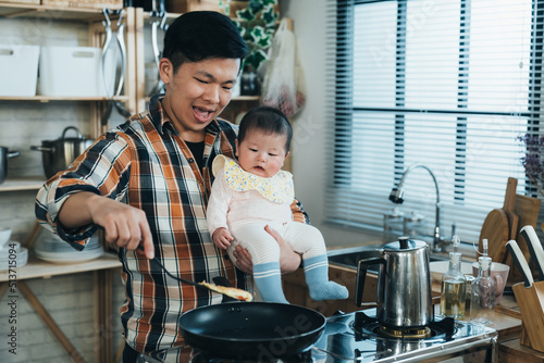 Obraz na plátně happy Korean father carrying his innocent baby girl in arm is using a spatula to fry food by the kitchen stove during daytime at home