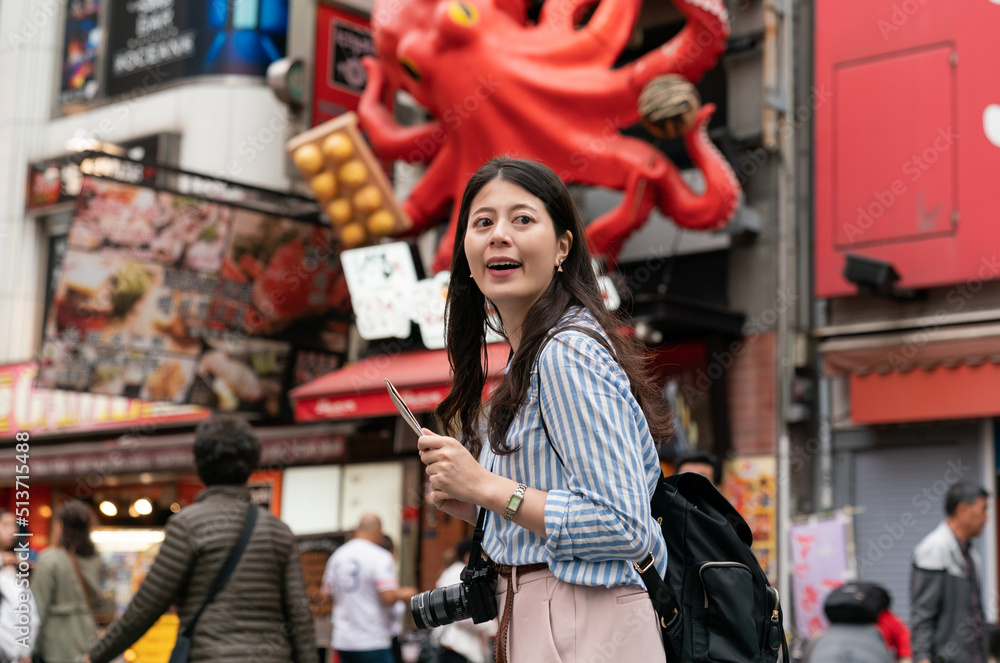 excited Asian woman visitor looking around with map in hand while enjoying active vibe at the shopping district in shinsaibashisuji and dotonboshiri Osaka japan with octopus sign behind