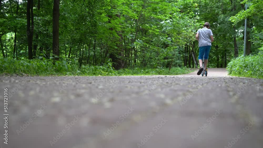 The boy is riding a scooter in the park. View from the back. Summer, road and green trees. Kick scooters drive fun The boy is riding a scooter in the park. View from the back. Summer, road and green trees. Kick scooters drive fun