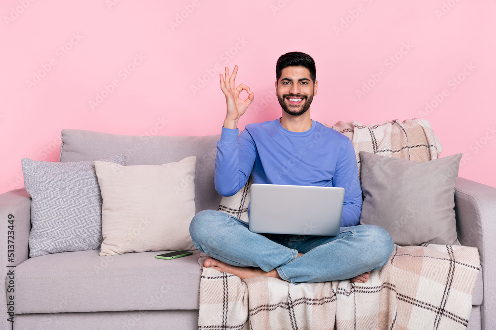 Photo of attractive confident arabian man wear long sleeve shirt sitting sofa modern gadget okey sign empty space isolated pink color background Photo of attractive confident arabian man wear long sleeve shirt sitting sofa modern gadget okey sign empty space isolated pink color background