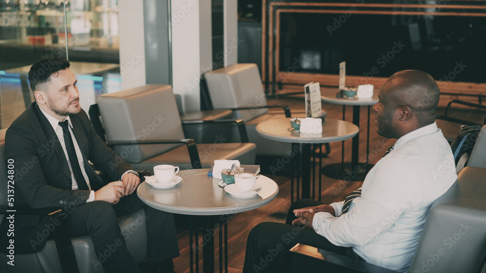 Two multi-ethnic businessmen sitting, smiling and discussing their startup in airy cafe during lunch time. Coffee cups are on their table Two multi-ethnic businessmen sitting, smiling and discussing their startup in airy cafe during lunch time. Coffee cups are on their table