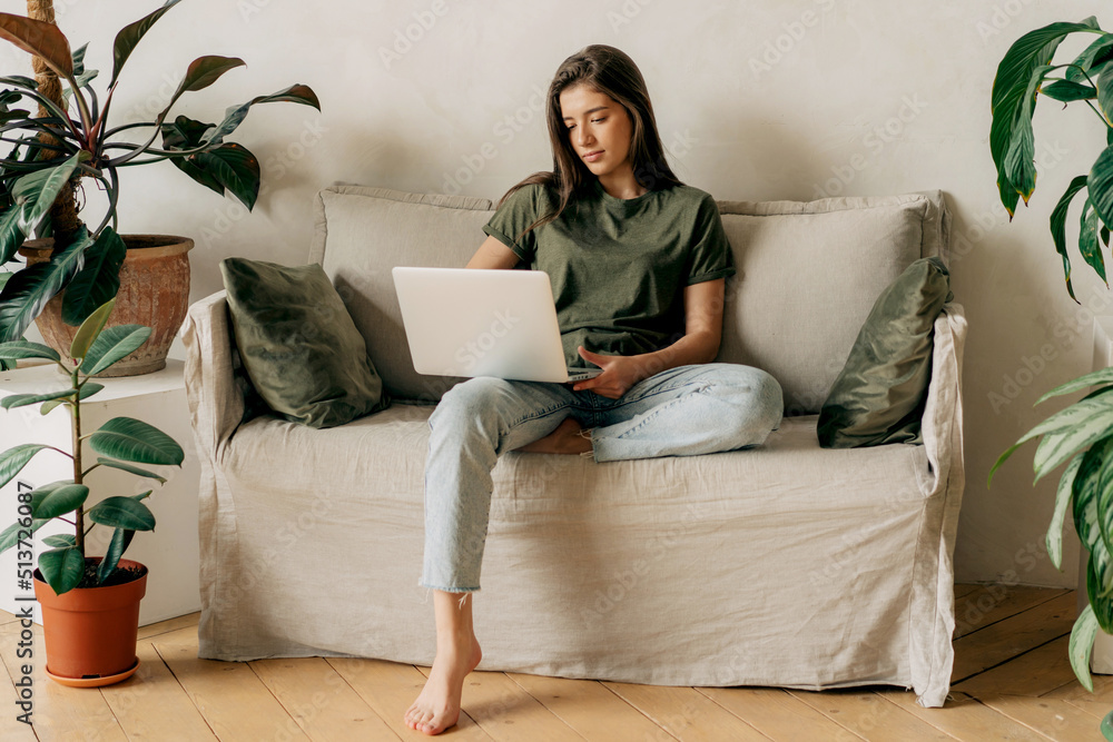 A young attractive brunette girl sitting on a sofa uses a laptop to work remotely online. A young attractive brunette girl sitting on a sofa uses a laptop to work remotely online.