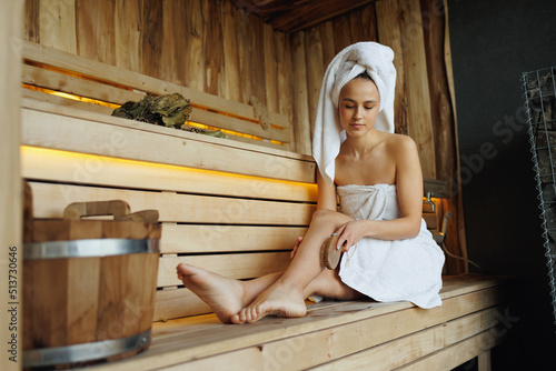 Beautiful woman in white bathrobe and towel on head doing body massage with dry wooden brush with natural bristles in sauna photo