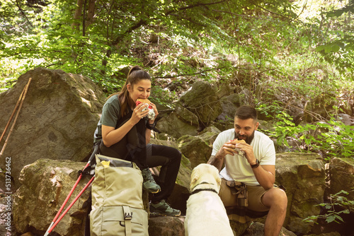 Young hikers couple sitting on the rocks on the mountain woods eating a sandwiches at break while their dog watch them. Nature lovers hiking through the forest eating lunch. With film grain