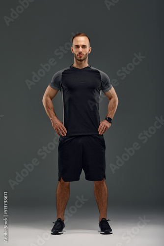 Full length portrait of young sporty guy in t-shirt and shorts standing on gray studio background. Serious millennial sportsman looking at camera. Healthy lifestyle and sports concept