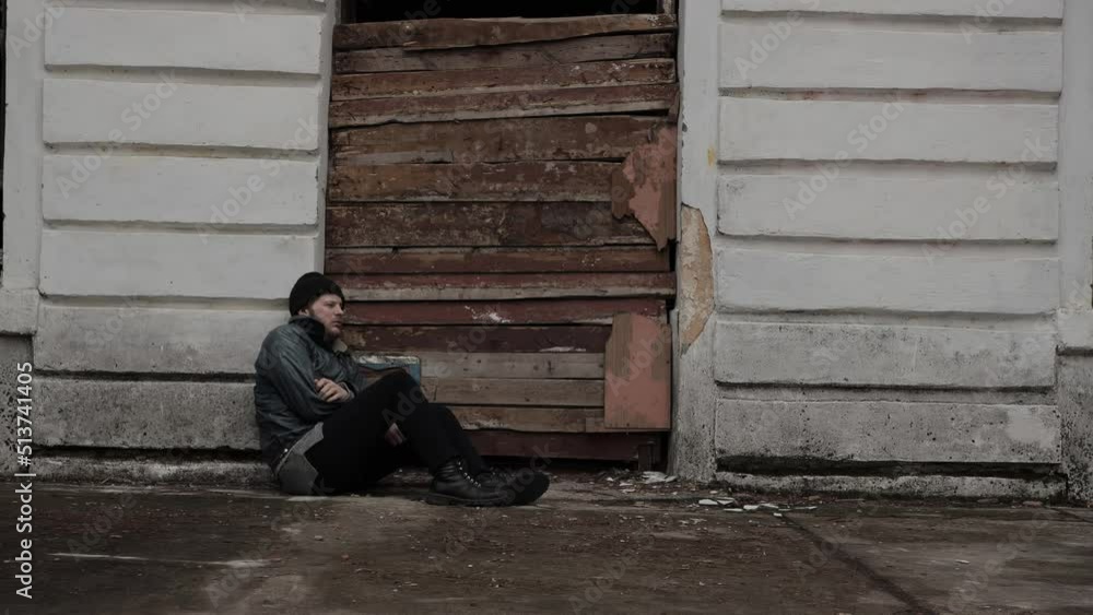 Homeless man sits outside an abandoned building. Refugee needs help. Below poverty line. Illegal immigrant. Tramp wearing in dirty clothes and hat cap. Fugitive. Consequences of drunkenness alcoholism Homeless man sits outside an abandoned building. Refugee needs help. Below poverty line. Illegal immigrant. Tramp wearing in dirty clothes and hat cap. Fugitive. Consequences of drunkenness alcoholism