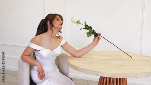 Beautiful girl sidiya at the table sniffing a white rose, posing. Charming young bride, brown hair, light isolated background, photo studio, copy space. Sparkling flash, soft light. UHD 4K.