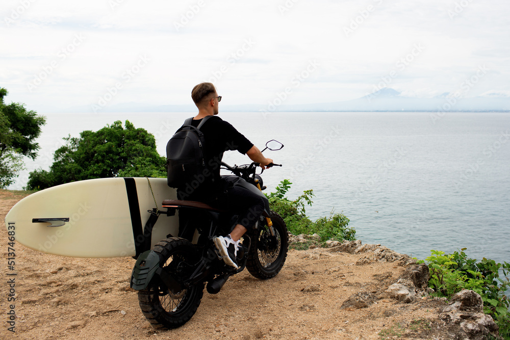 Fototapeta premium young man sits on a motorcycle with a surfboard.