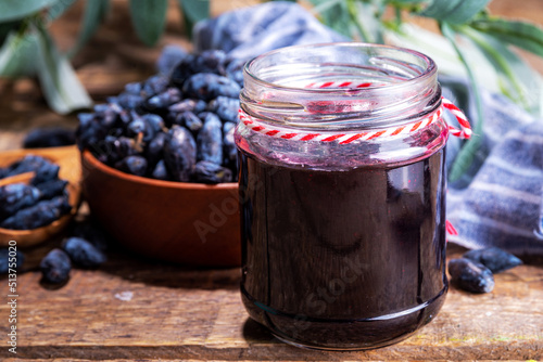 Small glass jar with homemade haskap berry jam, on wooden background with bowl of fresh haskap (honeysuckle, honeyberry) berries, copy space