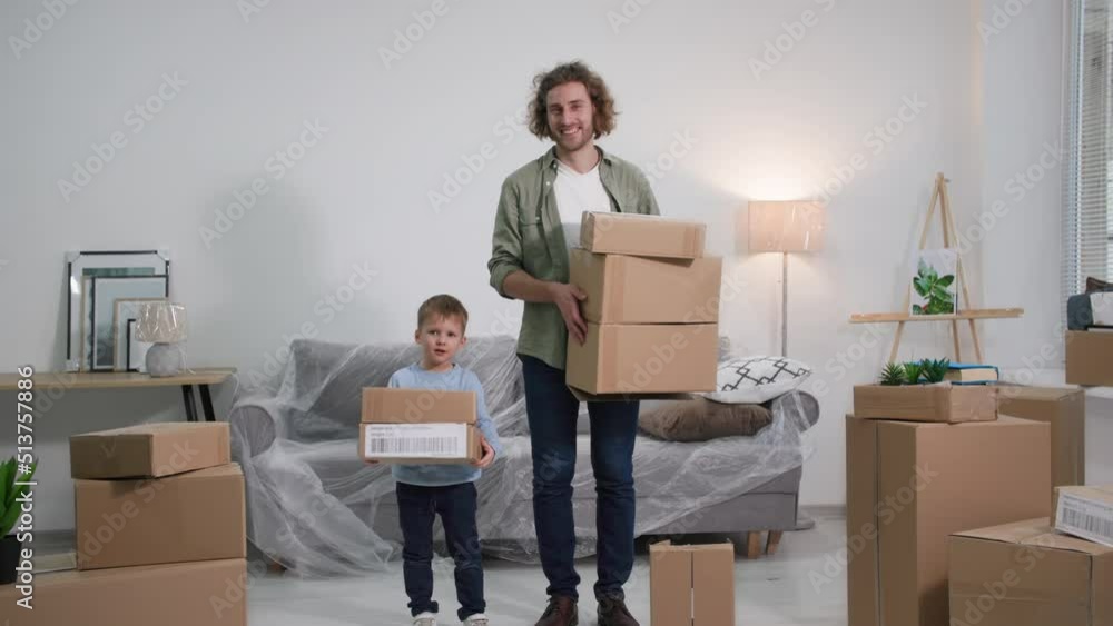 young handsome father and son stand with cardboard boxes in new apartment with furniture covered with polyethylene after the move, portrait of a parent and a boy during the rest young handsome father and son stand with cardboard boxes in new apartment with furniture covered with polyethylene after the move, portrait of a parent and a boy during the rest