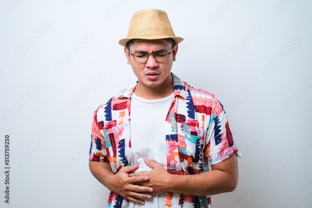 Young handsome Asian man wearing casual beach shirt and bucket hat diarrhea health problems hold his belly Young handsome Asian man wearing casual beach shirt and bucket hat diarrhea health problems hold his belly
