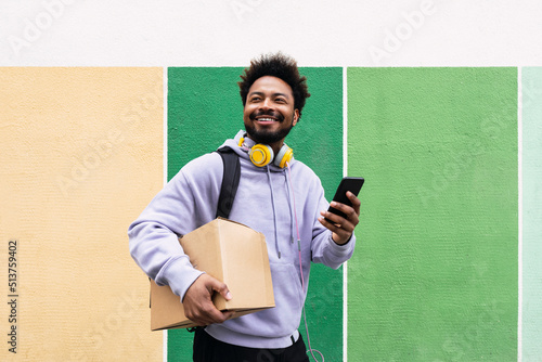Smiling delivery man holding box and mobile phone in front of colorful wall photo