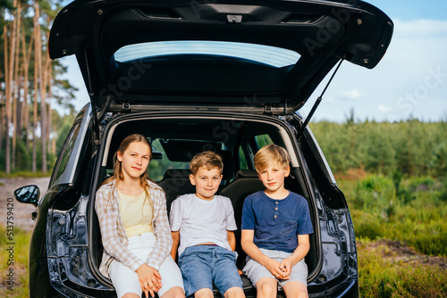 Three children siblings looking at camera sit at trunk of in car forest. Family goes to forest for weekend. summer travel trip. © Iryna