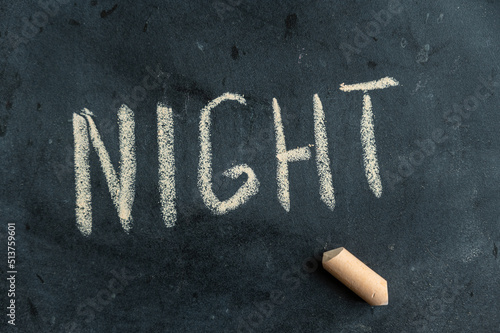 NIGHT. The name of the time of day is written in yellow chalk on a black chalkboard. The text is handwritten. Next to it is a piece of colored chalk.