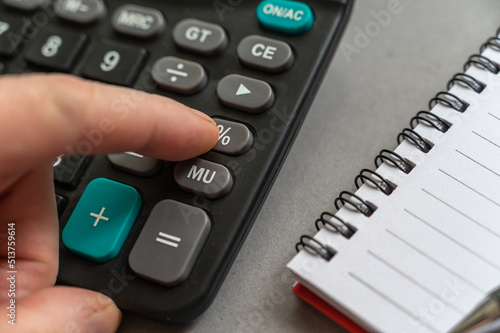 Close-up of male hand doing calculations on calculator. Index finger presses the percentage button. An open spring-loaded notebook and a pencil lie side by side on a gray surface. Selective focus.