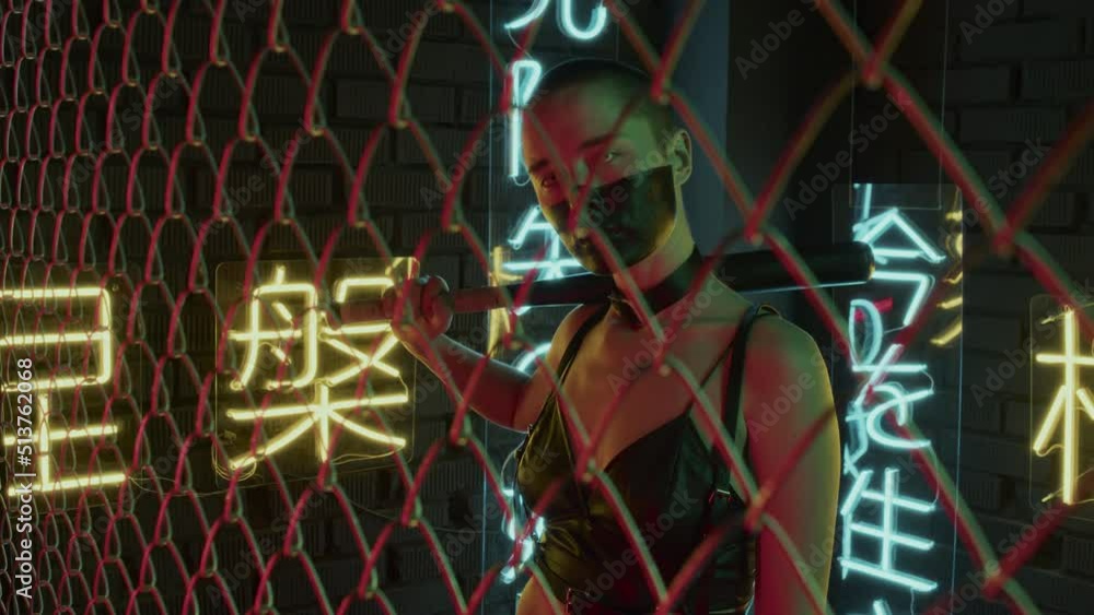 Waist-up of girl with shaved head, black paint on face, in white contact lenses hitting fencing with bat in studio with neon lights in shapes of hieroglyphs meaning nirvana and western paradise Waist-up of girl with shaved head, black paint on face, in white contact lenses hitting fencing with bat in studio with neon lights in shapes of hieroglyphs meaning nirvana and western paradise