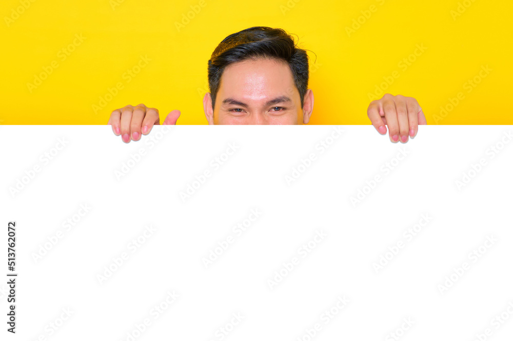 Smiling young Asian man in casual t-shirt holding hiding behind white advertisement board isolated on yellow background. Promotion billboard concept