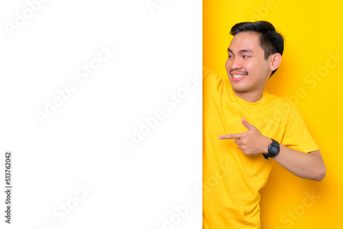 Cheerful young Asian man in casual t-shirt pointing at white advertisement board isolated on yellow background. Promotion billboard concept