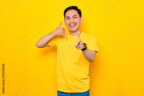 Cheerful young Asian man in casual t-shirt showing call me sign gesture and pointing at camera isolated on yellow background. People lifestyle concept © Sewupari Studio