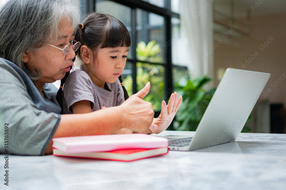 Asian Grandmother with her two grandchildren having fun and playing education games online with a computer notebook at home in the living room. Concept of online education and caring from parents. Asian Grandmother with her two grandchildren having fun and playing education games online with a computer notebook at home in the living room. Concept of online education and caring from parents.