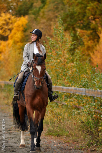 Young beautiful brunette girl rides a horse on a warm and sunny autumn day. Portrait of a pretty young woman on the horse, wearing tall boots and gloves. © Georgii