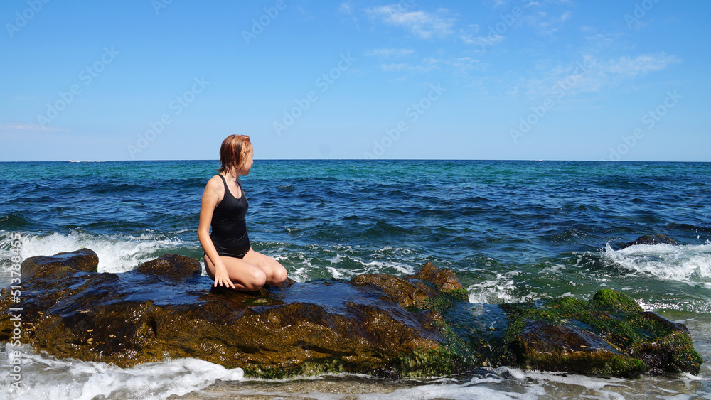 A teenage girl in a black swimsuit sitting on a large rock on the beach against the sea horizon