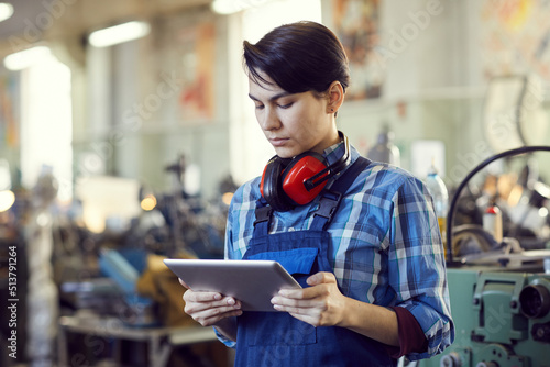 Serious busy female factory engineer with short black hair standing in workshop and using tablet for lathe examination